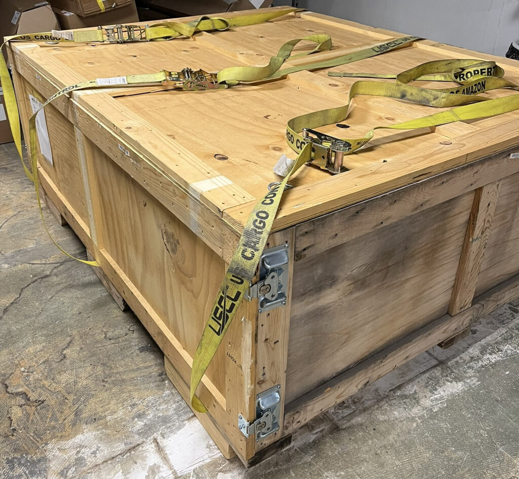 Figure 4: Large electric vehicle battery shipping crate stored at floor level with cargo straps and secured wood packaging. The image illustrates specialized battery packaging and handling considerations for warehouse storage and transport. Courtesy: Telgian Engineering & Consulting